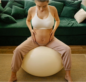 Grávida sentada em uma bola de pilates com as mãos na barriga.